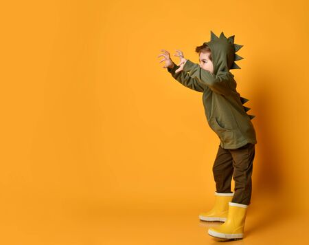 Cheerful kid boy in dark green hoodie with dinosaurs saurus spikes at his back and cowl playing t-rex dangerous beast with his hands claws up and roars at free copy space over backgroundの写真素材