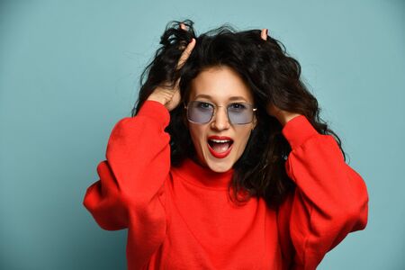 Much surprised woman in red sweater and stylish rectangular glasses hold her hands run her fingers through her long curly hair over light blueの写真素材