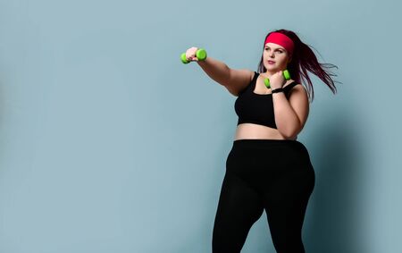 Fitness spring diet weight loss concept. Lucky plus-size girl overweight woman dieting working out with small green weights dumbbells in gym on pastel blue backgroundの写真素材