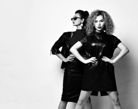 Black and white portrait of two young women in stylish black leather clothing dress, sjirt and jacket stand with hands on waist. Beauty, fashion, sexuality, trendy outfit conceptの写真素材