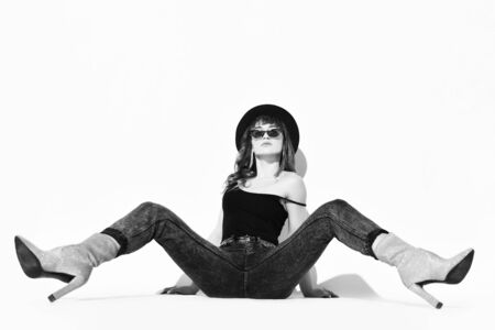 Young beautiful brunette woman in stylish casual clothing, hat and high heels boots sitting on floor with legs stretched out over white wall background. Stylish look and footwear conceptの写真素材