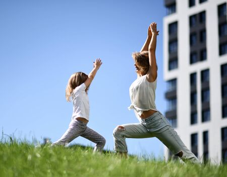 Mother and daughter together practicing yoga outside on a grass together urban city on blue sky backgroundの写真素材