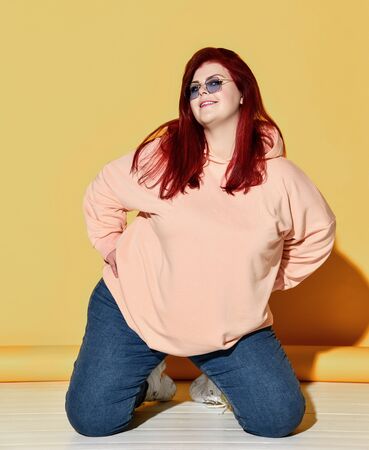 Young smiling red haired overweight woman in casual clothing, sneakers and sunglasses posing on floor over yellow wall background. Stylish look of pretty overweight womanの写真素材