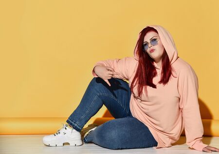 Young smiling red haired overweight woman in casual clothing, sneakers and sunglasses sitting on...
