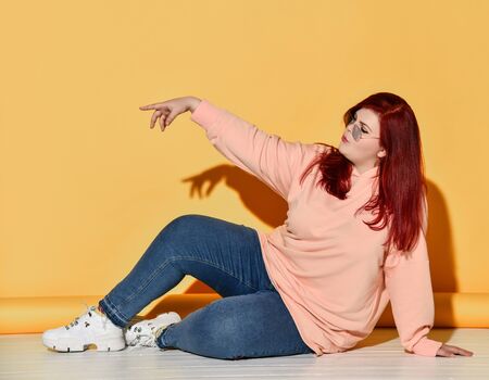 Young cheerful red haired plus size woman in casual clothing and sneakers sits on floor and feeling gorgeous commands on yellow background. Stylish look of pretty overweight womanの写真素材