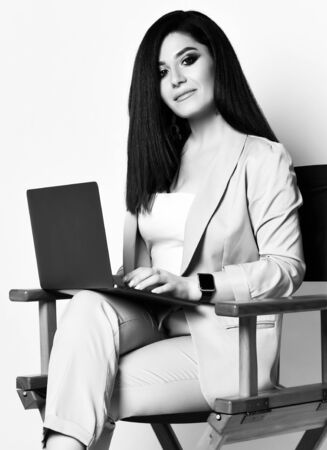 Black and white portrait of young self-confident business woman brunette in official pantsuit sitting on wooden armchair with black laptop on white backgroundの写真素材
