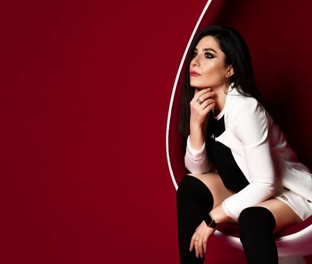 Young stylish woman brunette in black and whit clothes sitting in a modern oval chair and looks up on red background with free text copy spaceの写真素材