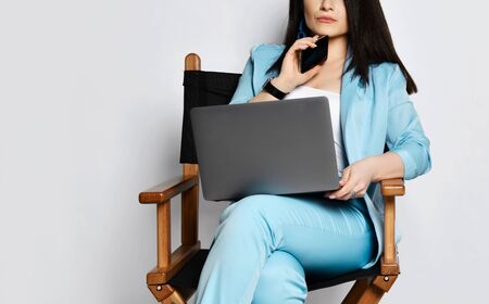 Portrait of young self-confident business woman brunette in blue official pantsuit sitting on wooden armchair with black laptop and cell phone at her chin on white backgroundの写真素材