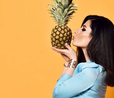 Young woman brunette with long straight hair in blue official wear jacket kisses a pineapple on yellow background with free text copy spaceの写真素材