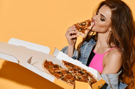 Portrait of sexy brunette woman in pink swimsuit and denim jacket holds a box of freshly baked tasty pizza eating slice on yellow background. Losing weight and getting fat conceptの写真素材