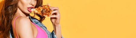Close-up of young curly brunette woman with pouty lips wearing pink bra and denim jacket eating pizza over yellow background. Sexy fit womans body and fast food concept. Bannerの写真素材