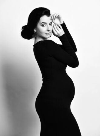 Black and white portrait of beautiful sensual pregnant woman posing side to us in tight dress holding hands at head, looking at camera. Happy maternity and beauty of pregnancy conceptの写真素材