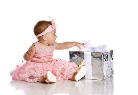 Infant child baby girl toddler sits with silver present gift box looking at it touching bow in pink dress celebrating birthday over white background with free copy spaceの写真素材