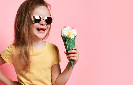 Happy smiling laughing kid girl in yellow t-shirt and sunglasses holds big vanilla ice cream with candies in waffles cone over pink on pastel pink background with copy spaceの写真素材