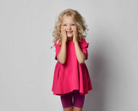 Beautiful positive little blonde girl with curly hair in stylish casual shorts and t-shirt holding her cheeks and smiling over grey wall background. Fashion for children conceptの写真素材