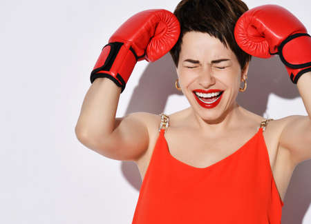 Happy screaming brunette woman with short hair in red dress, earrings and boxing gloves holds hand...
