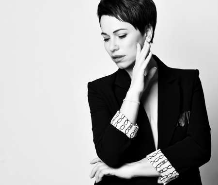 Portrait of romantic sad young short haired brunette woman in elegant business suit with deep neckline looking down holding hand at cheek over white background. Business elegance conceptの写真素材