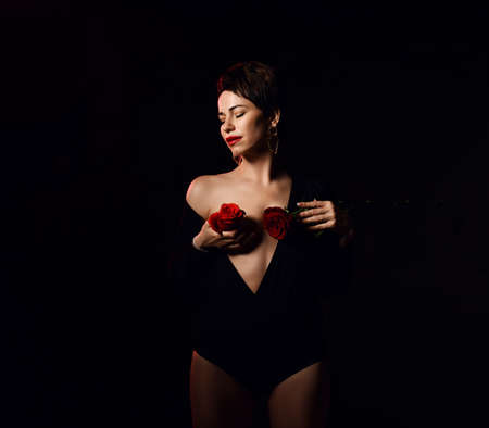 Young beautiful short haired brunette woman in undone bodysuit with naked breast holding red rose flowers over chest with eyes closed over dark background. Sexy woman conceptの写真素材