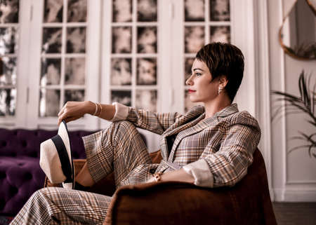 Positive and calm short haired brunette woman in stylish business plaid pantsuit with rolled up sleeves sits in armchair  holding hat in hand in room. Business elegance conceptの写真素材