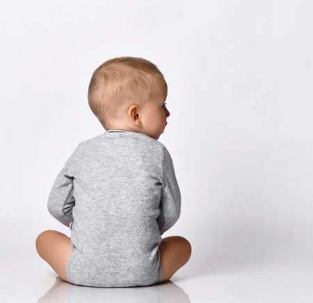 Toddler one-year-old baby boy in diaper and grey one-piece bodysuit with long sleeves sits back to camera looking aside with interest reading. Happy infancy and babyhood conceptの写真素材
