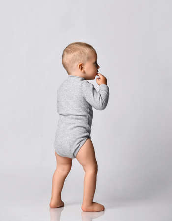 Toddler one-year-old baby boy in grey one-piece bodysuit with long sleeves stand back to camera looking aside holding hand at his mouth. Happy infancy and babyhood conceptの写真素材