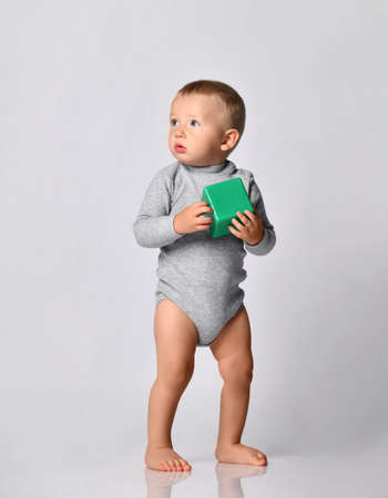 Toddler one-year-old baby boy in diaper and grey one-piece bodysuit with long sleeves stands holding green toy block looking at upper corner. Happy infancy and babyhood conceptの写真素材
