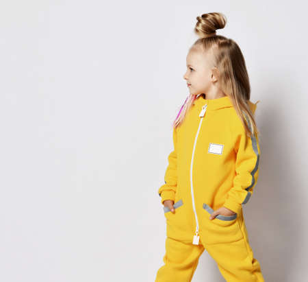 Blonde kid girl with modern hairstyle and braids in stylish yellow jumpsuit stands with her hands in pockets and looks aside at copy space over gray backgroundの写真素材