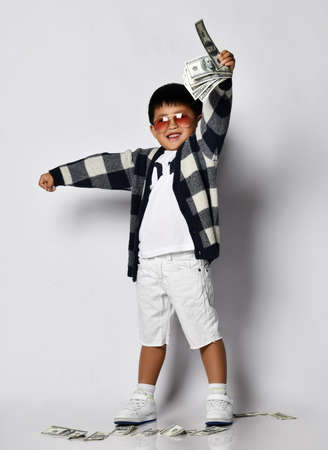 Happy smiling asian rich kid boy millionaire in modern sunglasses raised his hand with dollars up cash bunch of money. Lucky winner. Save money. Overspend. White backgroundの写真素材