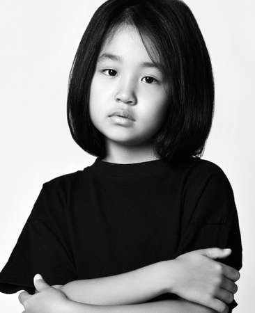 Black and white portrait of asian kid girl in black t-shirt and bob hairstyle standing with her arms crossed at chest and looking at camera with her head tiltedの写真素材