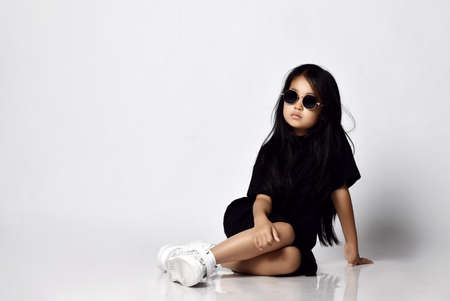 Pretty serious brunette chinese girl in stylish black dress, white sneakers and sunglasses sitting on floor and looking aside over white wall background. Trendy children fashion conceptの写真素材