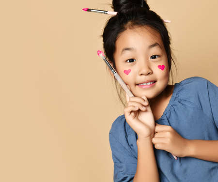 Portrait of smiling pretty asian girl in blue cotton dress with painted hearts on cheeks with brush...