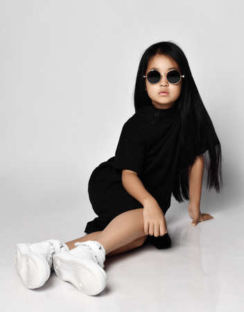 Serious self-confident asian kid girl in stylish black dress, white sneakers and round sunglasses...