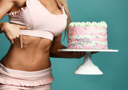 Hands of sexy pretty fitness slim woman in pink bra holding birthday cake with cream and pointing at slim sporty belly with fingers over blue background, cropped. Figure, present, sport, holidayの写真素材