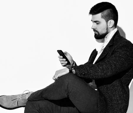 Young brunette serious bearded brutal businessman in stylish official costume and boots sitting and...