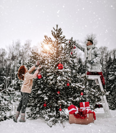 Young happy couple in stylish winter clothing decorating Christmas tree with balls outdoors in winter snowy forestの写真素材