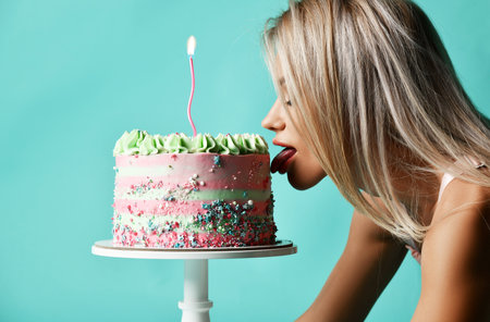 Portrait of young sexy blonde woman standing on all fours and licking big cake with cream and candle. Side viewの写真素材