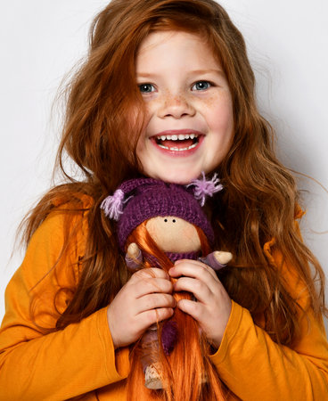 Little five-year-old red-haired girl smiles happily and holds a redhair doll in her hands in an orange sweater on whiteの写真素材