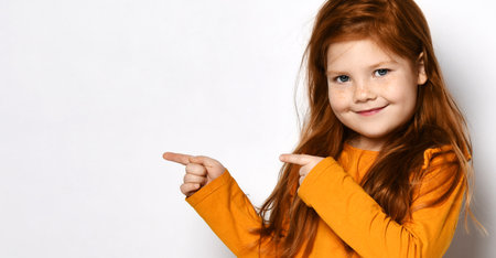 Smiling small red haired girl with freckles in yellow comfortable longsleeve standing and pointing...