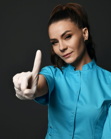 Young positive woman doctor in blue working uniform and latex gloves showing forefinger to cameraの写真素材