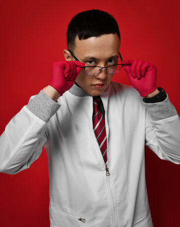 Portrait of young man doctor urologist or proctologist in medical gown and gloves putting on taking off his glassesの写真素材