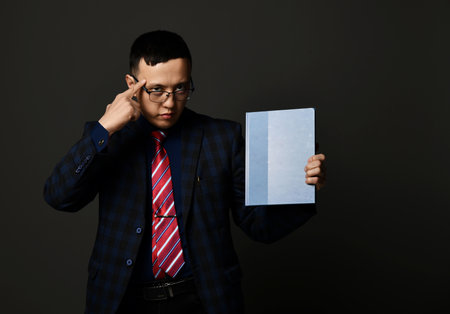 Young asian serious businessman standing holding blank blue paper in hand and pointing at head...