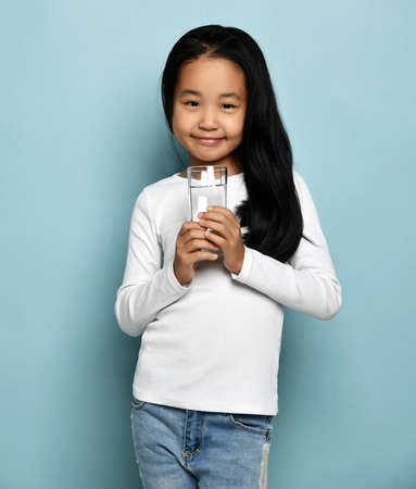Cute smiling asian Korean kid girl in jeans and white long sleeve shirt stands holding full glass of pure waterの写真素材