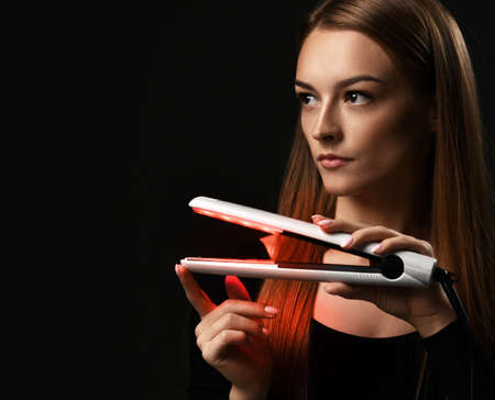 Portrait of young refined woman holding modern hair straightener in hands and looking aside at copy...