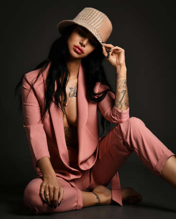 Sexy brunette woman with tattoo on body arms and legs wearing pink silky pantsuit on naked body and...