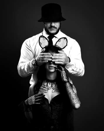 Black and white. Woman with tattoo in lingerie and sexy rabbit mask sits with man in suit behind her covering her eyesの写真素材