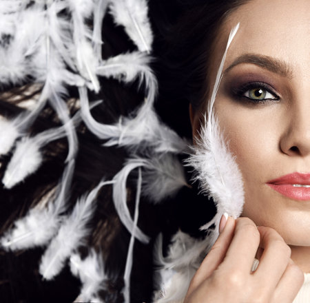 Half-face portrait of graceful gorgeous brunette woman surrounded with falling white feather and fluffの写真素材