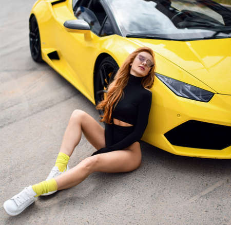 Hot sexy sporty red-haired woman in bodysuit and naked legs, long gloves and sunglasses sits on asphalt at sport carの写真素材