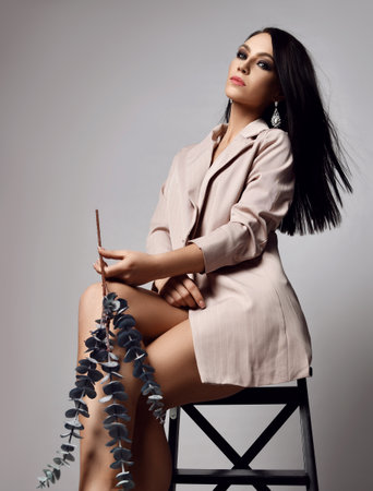 Pretty brunette woman in beige jacket sits on stepladder with dried exotic flower in hand looking down at cameraの写真素材