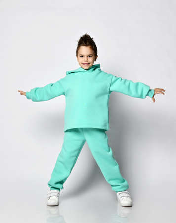 Smiling active kid boy with stylish haircut in modern green, mint color sportswear hoodie stands holding hands up spreadの写真素材