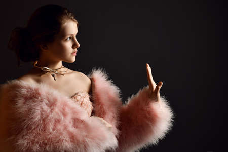 Portrait of kid girl, young lady in evening dress and fur coat with big sleeves holding hand up and looking asideの写真素材